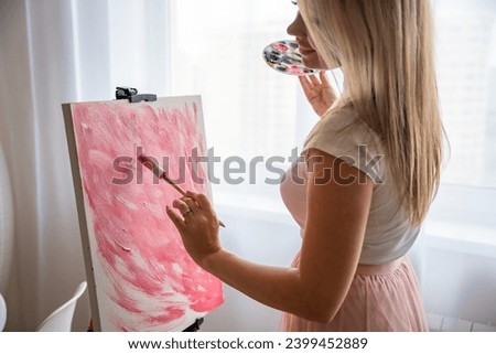 Young blonde woman artist with palette and brush painting abstract pink picture on canvas at home. Art and creativity concept. High quality photo