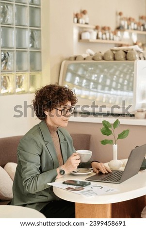 Young female entrepreneur looking through information on laptop screen while having coffee at cafe