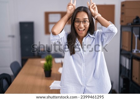 Young hispanic woman at the office posing funny and crazy with fingers on head as bunny ears, smiling cheerful 