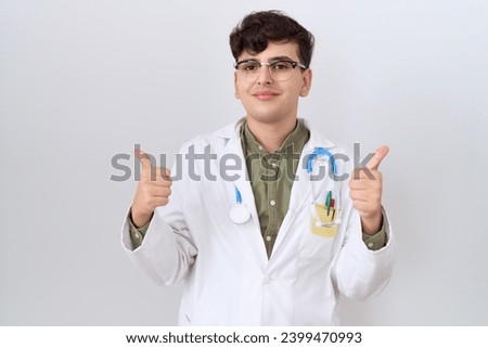 Young non binary man wearing doctor uniform and stethoscope success sign doing positive gesture with hand, thumbs up smiling and happy. cheerful expression and winner gesture. 