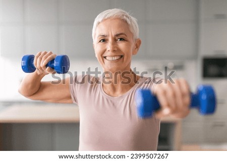 Home workout. Happy mature woman exercising with dumbbells smiling to camera indoors, portrait of healthy fitness lady training in domestic interior, keeping fit, leading active sporty lifestyle Royalty-Free Stock Photo #2399507267