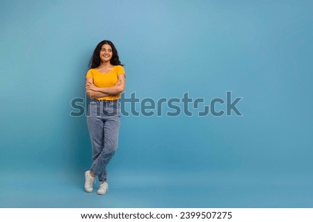 Cute positive pretty curly young indian woman in casual clothing holding hands crossed on chest, looking at copy space for advertisement and smiling, blue studio background, full length Royalty-Free Stock Photo #2399507275