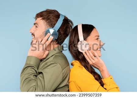 Cheerful young man in green shirt and woman in yellow blouse enjoying music on headphones while standing back to back against blue background, side view