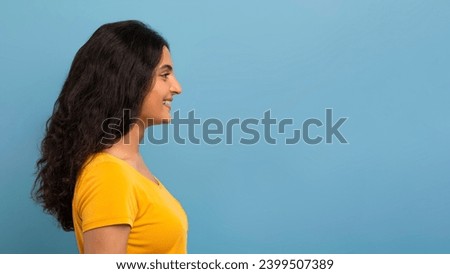 Profile studio shot of happy curly young indian woman student in yellow t-shirt looking at blank copy space for text or advertisement and smiling, blue background, panorama Royalty-Free Stock Photo #2399507389