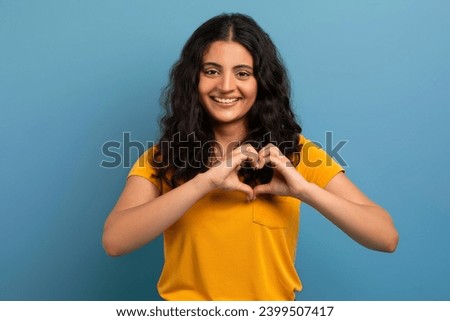 Cute cheerful beautiful curly young indian woman student wearing yellow t-shirt showing heart gesture over chest and smiling at camera, expressing love and gratitude over blue background Royalty-Free Stock Photo #2399507417