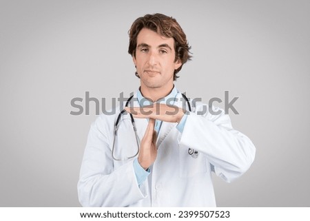 Confident young man in white coat with stethoscope doing time out gesture with hands, standing isolated on grey studio background. Stop sign, time to break