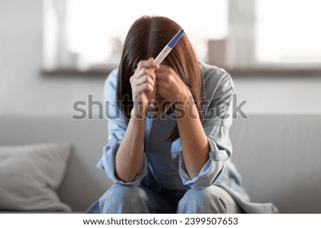 Unrecognizable young woman holding pregnancy test with negative result, covering face in despair sitting at home. Maternal health problems and infertility challenges concept Royalty-Free Stock Photo #2399507653