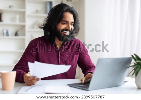 Handsome Indian freelancer guy working on his laptop and checking documents, smiling eastern man sitting at desk in home office, organizing paperwork and using computer, enjoying remote work