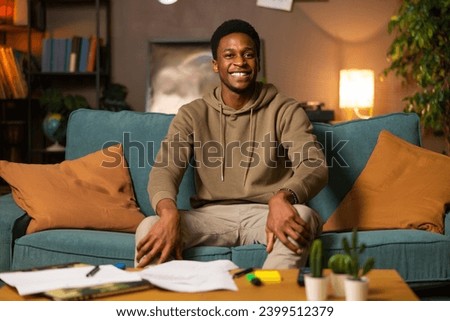 Photo of delighted dark skinned african american black man guy student sitting in the living room smiling having mess at home studying educational proess online from distance preparing for exams.
