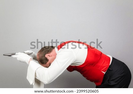 Portrait of a Waiter or Servant in Red Vest Bowing Profusely and Offering Silver Tray. Act of Sycophantic Servitude. Royalty-Free Stock Photo #2399520605
