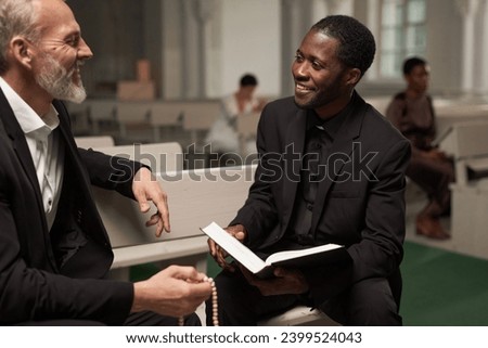 Portrait of young Black man as smiling priest talking to senior man after Sunday service in catholic church, copy space Royalty-Free Stock Photo #2399524043