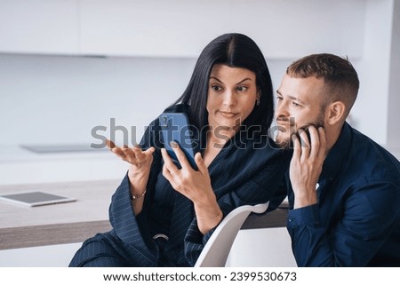 Surprised couple looking at phone reading unpleasant message at home. Young woman in suit shows to husband news on phone via internet. Unhappy young man watching awful videos. Royalty-Free Stock Photo #2399530673