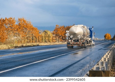 Commercial blue big rig semi truck tractor transporting liquid cargo in tank semi trailer running on the autumn highway road with wet reflective after the rain surface and red maples on the side Royalty-Free Stock Photo #2399542605