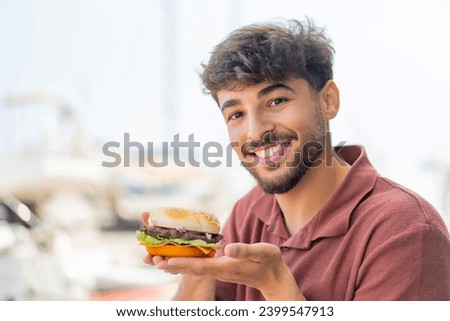 Young Arabian handsome man at outdoors holding a burger with happy expression