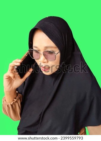 an adult Asian Muslim woman in a black hijab, brown blouse and glasses uses a smartphone to communicate, on a green background. communication concept