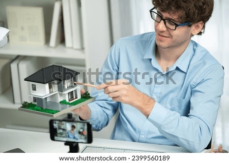Host channel of architecture using house model to teach architect by smartphone record in small model, pointing to explain modern home design communicating listeners on social media at home. Gusher.