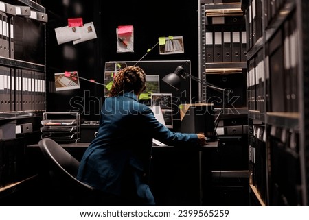 African american detective reviewing clues to solve crime case. Police criminologist woman standing near workplace desk, looking at evidences scheme and searching insight at night time
