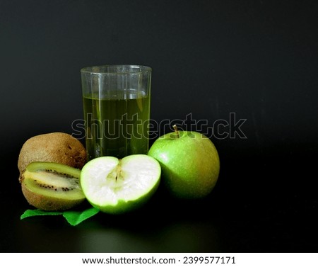 A tall glass of green fruit juice on a black background, next to pieces of a ripe green apple and kiwi with leaves. Close-up.
