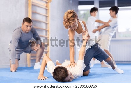 Focused teenager learning effective self defence techniques in sparring with boy under guidance of experienced instructor, practicing painful armlock on opponent lying face down on floor in gym.. Royalty-Free Stock Photo #2399580969