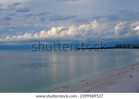 from above and from a distance, a small number of people enjoy the sandy beach and ocean water while a large line of billowy white clouds build up at the horizon near buildings Royalty-Free Stock Photo #2399589527