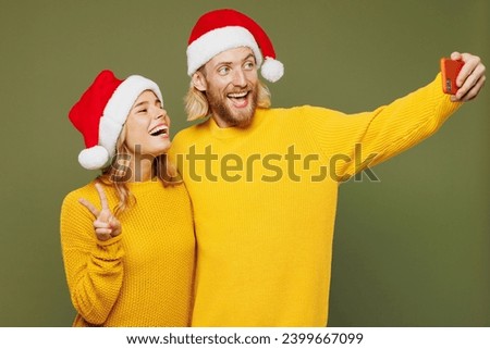 Merry young couple two friends man woman wears sweater Santa hat posing doing selfie shot on mobile cell phone show v-sign isolated on plain green background. Happy New Year Christmas holiday concept