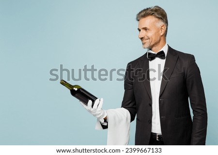 Side view adult sommelier barista male waiter butler man wears shirt black suit bow tie uniform hold in hand bottle of wine work at cafe isolated on plain blue background. Restaurant employee concept Royalty-Free Stock Photo #2399667133