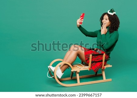 Full body merry little kid teen girl wear hat casual clothes posing sledding doing selfie shot on mobile cell phone isolated on plain green background studio. Happy New Year Christmas holiday concept