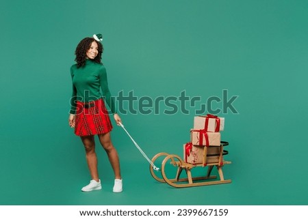 Full body fun merry little kid teen girl wear hat casual clothes posing carry on sled stack of gift present box isolated on plain green background. Happy New Year celebration Christmas holiday concept