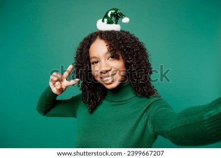 Merry little kid teen girl wear turtleneck hat casual clothes posing do selfie shot on mobile cell phone show v-sign isolated on plain green background studio portrait. Happy New Year holiday concept