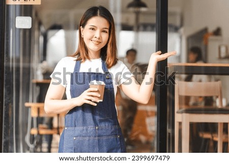 Happy excited attractive young Asian woman in denim apron, received online order in coffee shop with an 'Open' sign.