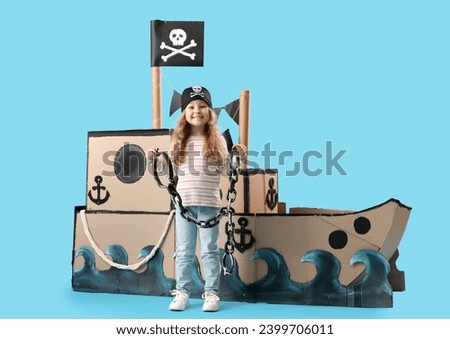 Cute little girl dressed as pirate with chain and cardboard ship on blue background