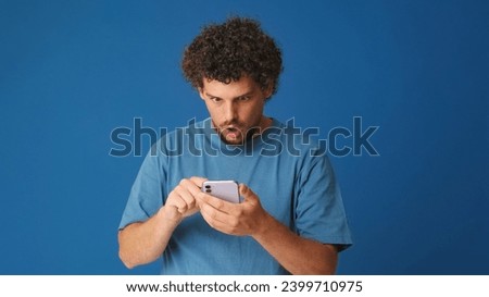 Amazed guy with curly hair dressed in blue t-shirt, holding mobile phone, shopping online, wow effect on blue background in the studio