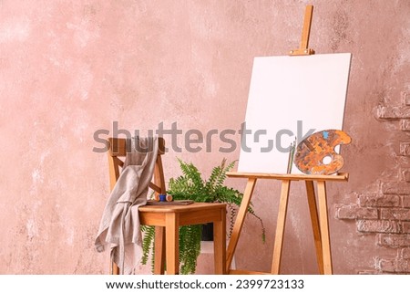 Wooden easel with blank canvas and painting tools near color wall in artist's workshop Royalty-Free Stock Photo #2399723133