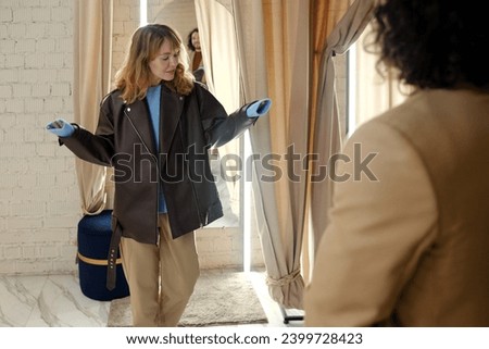 Young blond woman in stylish black leather jacket standing by fitting room in front of her husband while choosing new clothes in boutique Royalty-Free Stock Photo #2399728423