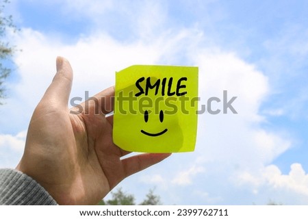 Hand holding note writing SMILE smile emoji with blue sky and clouds background. Motivational word wallpaper SMILE written on sticky note