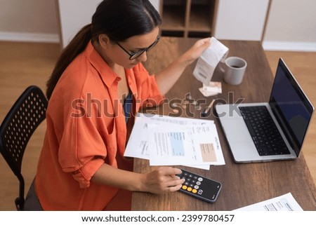 top view Desk Finance: A woman, with a laptop on her desk, tracks household expenses using her smartphone calculator. Royalty-Free Stock Photo #2399780457