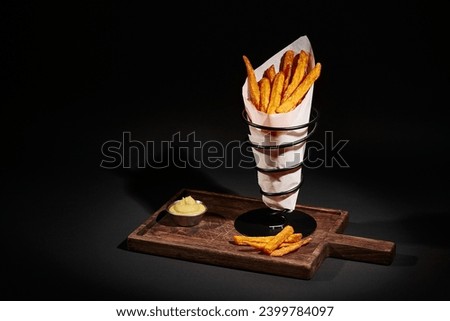 gourmet crispy French fries inside of paper cone near dipping sauce on wooden cutting board