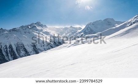 A serene, snow-blanketed mountainous terrain bathed in sunlight, with clear blue skies above and rugged peaks in the distance, suggesting a remote alpine region. Royalty-Free Stock Photo #2399784529