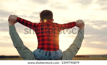Father carries preschooler son on shoulders supporting focused boy with hands. Dad and son enjoy tranquil walk deepening bonds spending time in field. Father with serene son creating memorable moment Royalty-Free Stock Photo #2399790879