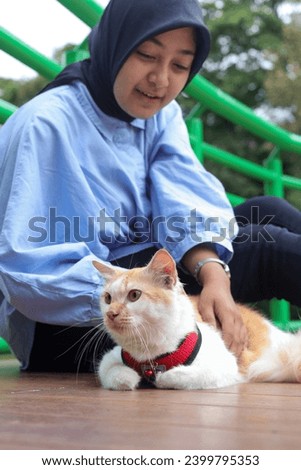 Outdoor portrait of beautiful Asian hijab woman holding and giving gentle touch to cat, taking care of her pet in nature park. Love relationship between humans and animals.