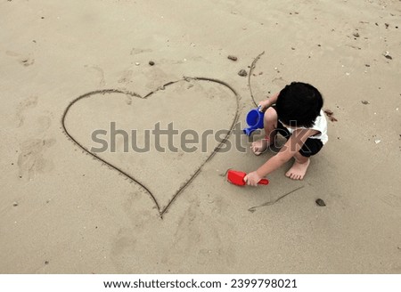 Exterior photo view of a little young 4 years old kid child boy who just draws traces of digs an big large heart shape to show his love to his mom dad parents at the beach during summer family holiday