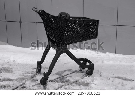 A black new plastic empty designer grocery cart stands in a snow-covered store driveway against a gray ventilated facade.