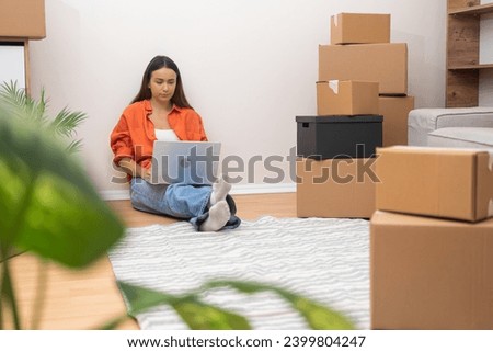 Energetic young woman makes their new apartment feel like home, laptop in hand. Boxes and sleek furnishings infuse the space with vibrancy