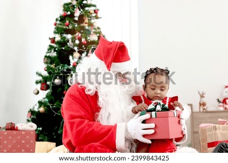 Happy smiling African child girl baby sitting on Santa Claus lap with decorative Christmas tree as background, cute kid open Christmas gift box present, feeling surprised and excited. Happy childhood