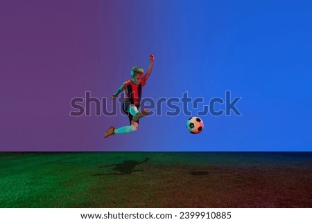 Active, energetic school age boy, football player training with soccer ball over dark blue background in neon. Concept of kids education, hobbies, skills, competition, achievement
