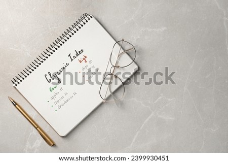 List with products of low and high glycemic index in notebook, pen and glasses on light grey marble table, top view. Space for text