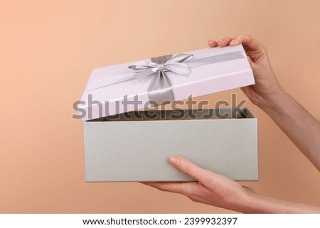 Side view of female hands opening gray purple box with satin ribbon and bow. Gift is hidden inside on a peach beige background. Open or close surprise color of the year 2024