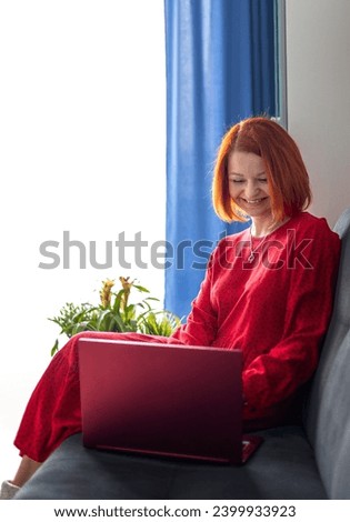  young woman in a red dress sits on a couch and looks laughing into a laptop camera. Video link