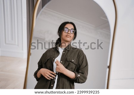 Young African American lady in casual shirt standing at mirror at living room looks away with pensive face dreaming about happiness, wealthy life.Girl in glasses thinks about better life. Royalty-Free Stock Photo #2399939235