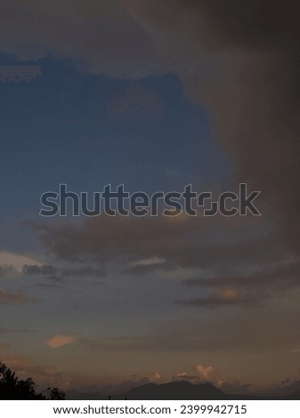 a sunset in a blue sky picture with large grayish white clouds, mountains in the background, tree silhouettes 